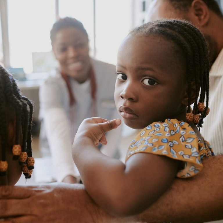 Portrait of young girl held in adult's arms while looking at camera in clinic setting with caregivers visible in background, displaying calm and serene expression